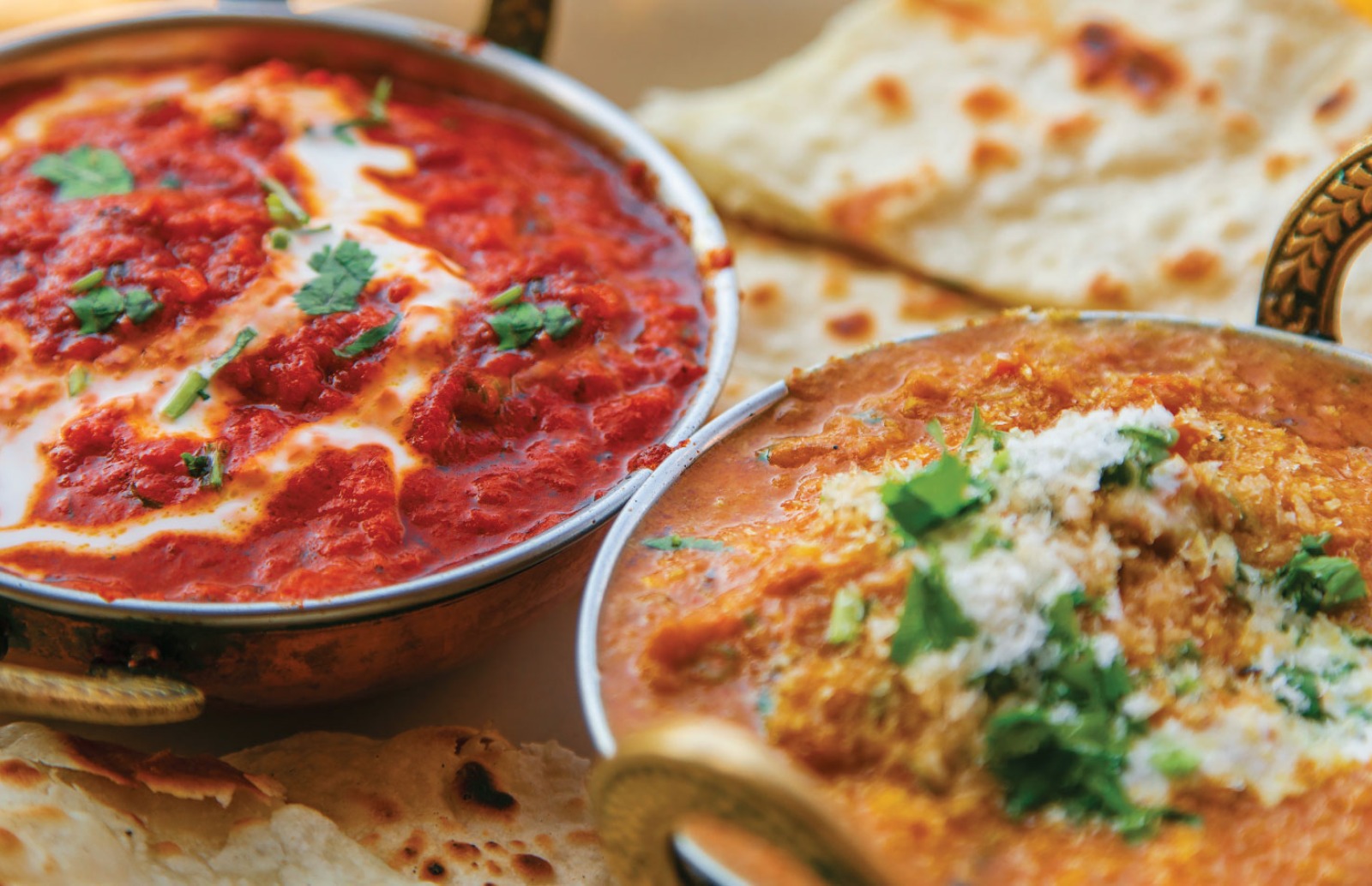 Two balti bowls filled with different curries. Garnished with coriander. Naan bread resting alongside the bowls